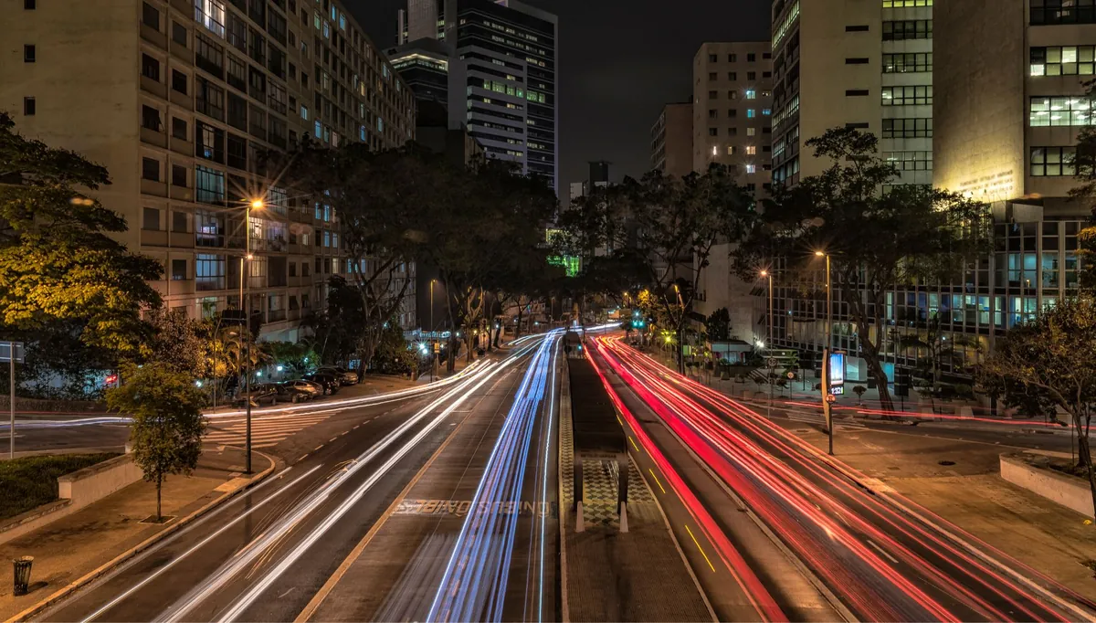 Avenida movimentada à noite com rastros de luz dos veículos entre prédios